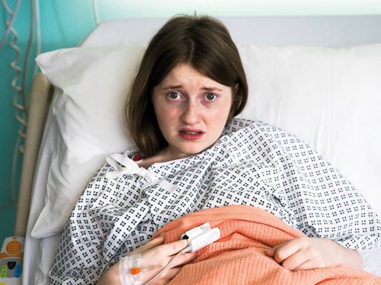 Auf dem Foto ist eine junge Person zu sehen, die in einem Krankenhausnachthemd in einem Krankenhausbett liegt. Sie schaut sehr verwirrt.